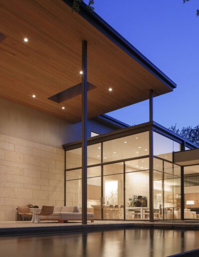 Modern house with large windows at dusk, featuring an illuminated interior and a pool in the foreground.
