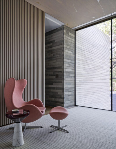 Modern interior with a pink wingback chair, matching stool, and a side table, featuring textured walls and floor-to-ceiling windows.