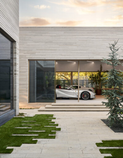 A modern car parked inside a contemporary house with large glass doors, surrounded by a neatly landscaped yard.