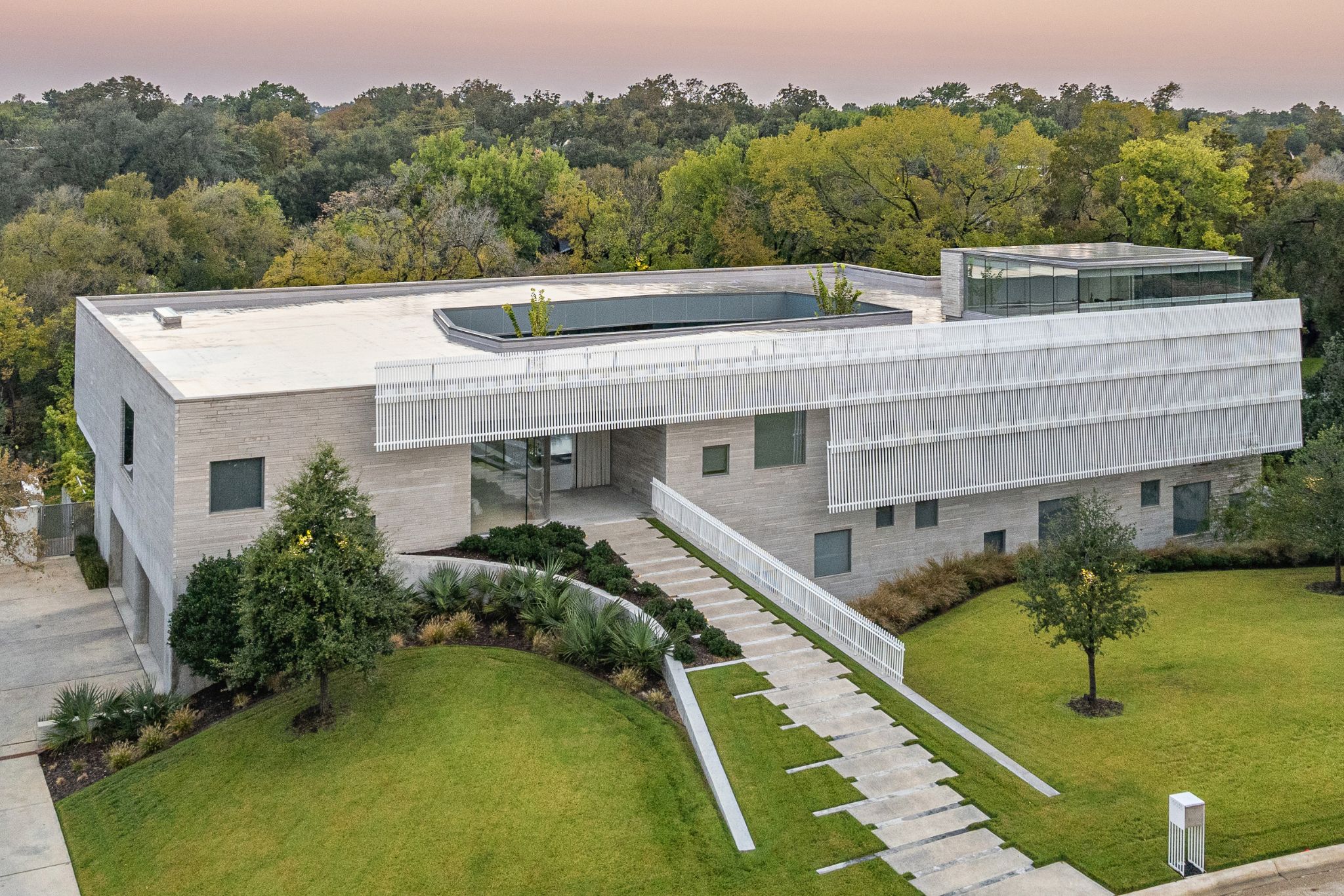 Modern white building with geometric design, large windows, and a flat roof. Surrounded by green lawn and trees. Concrete pathway leads to the entrance.