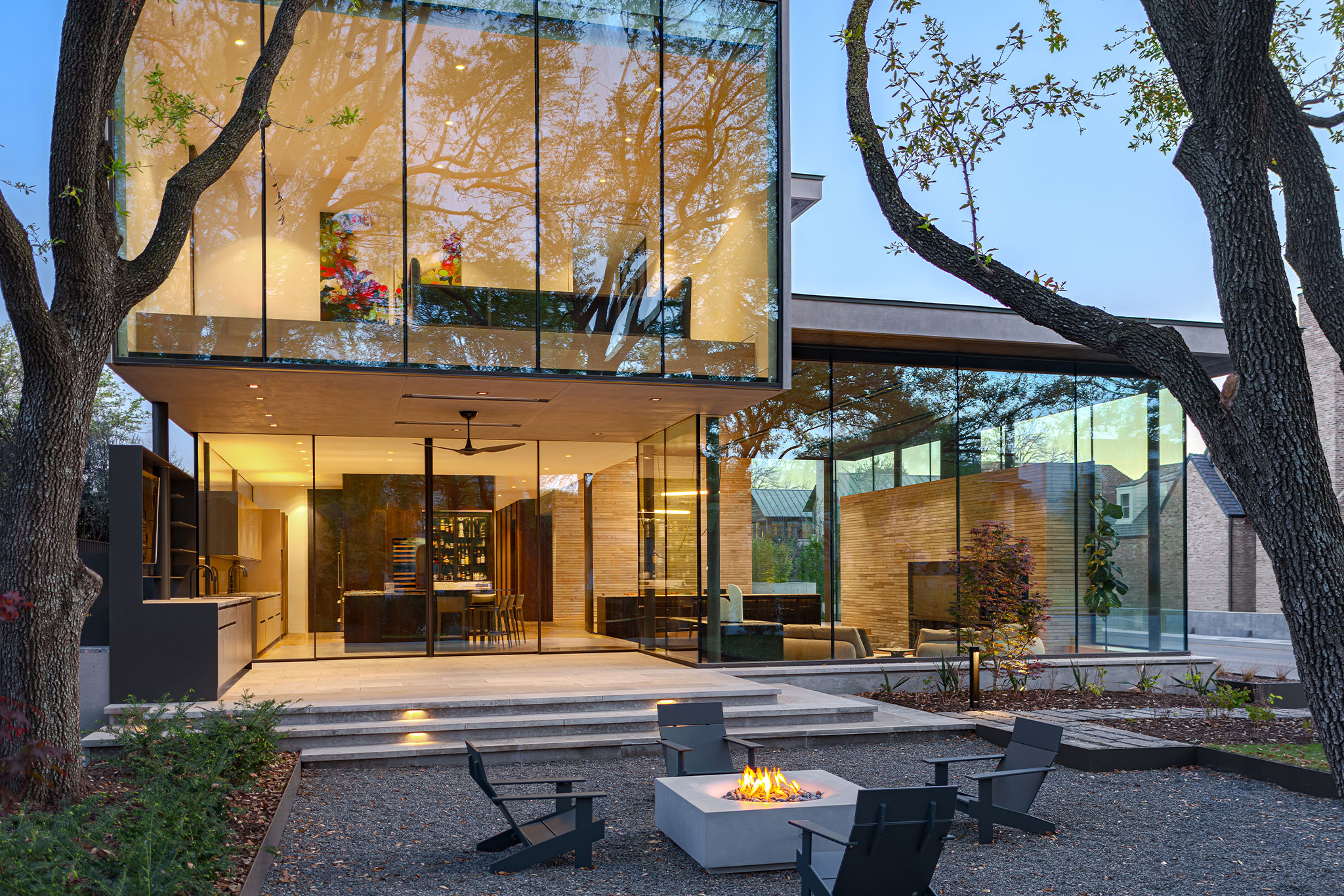 Modern house with large glass windows, surrounded by trees. A lit fire pit is in the foreground with black chairs arranged around it on a gravel patio.