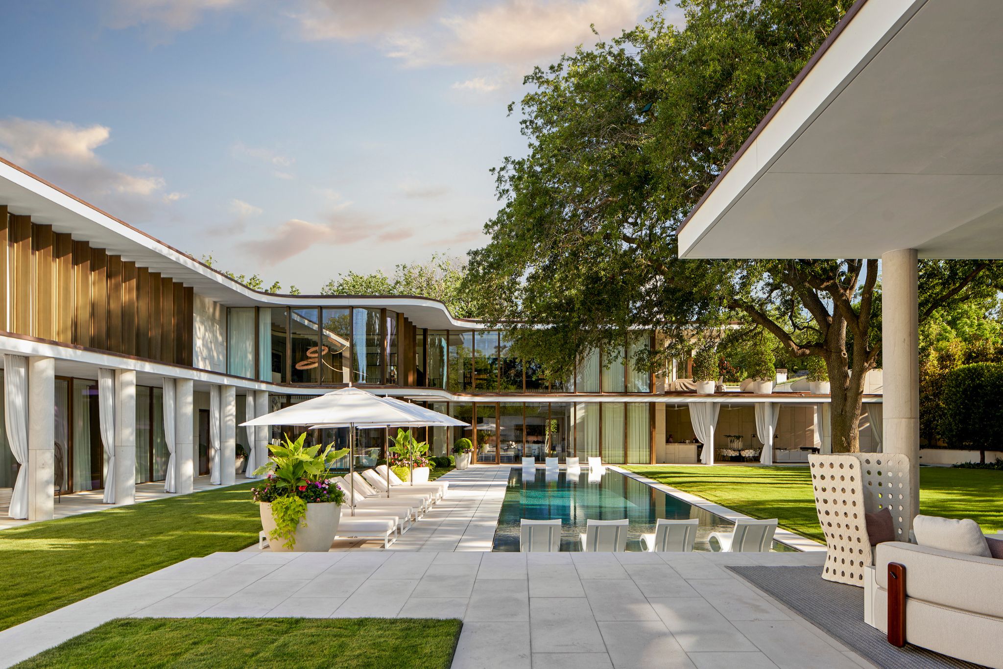 Modern house with large windows surrounding a rectangular pool, patio seating, and green lawn under a clear sky.